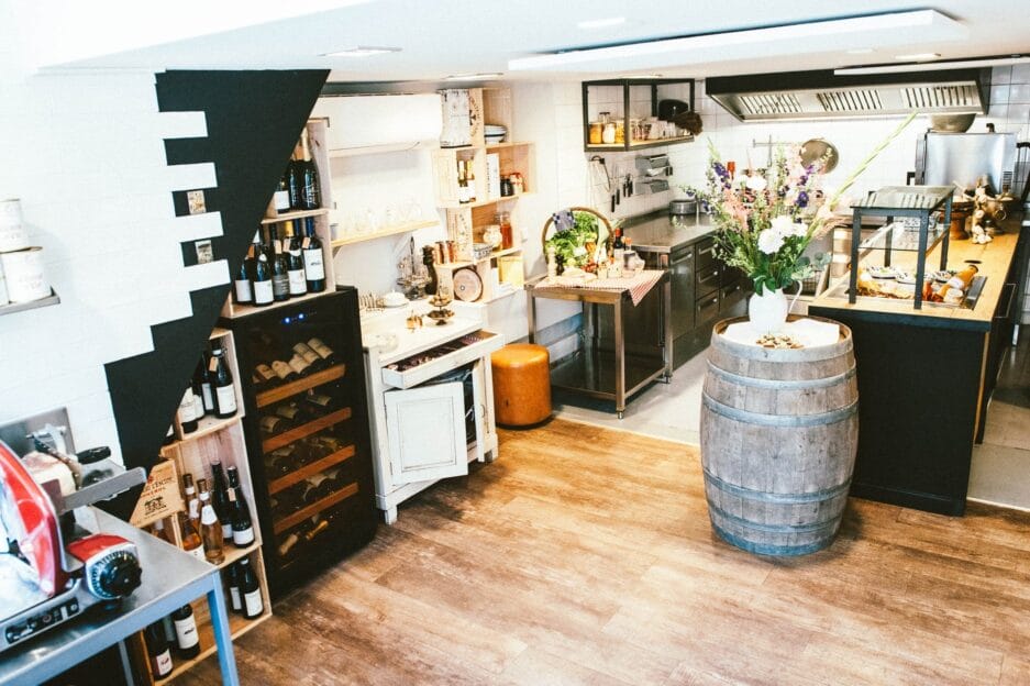 Interieur van een gezellige, rustieke restaurantkeuken met een houten vattafel, diverse flessen en een wijnbewaarkast.