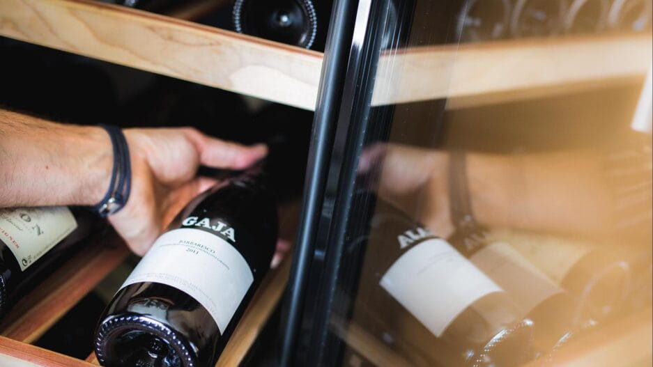 Een persoon die een fles wijn selecteert uit een koelkast gevuld met verschillende wijnen, zichtbaar door de glazen deur.