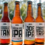 Vier flessen ambachtelijk bier met het opschrift 'goggle tan ipa', 'pocalypse ipa', 'joe ipa' en 'coast trip ipa' op een houten tafel buiten met vage bomen op de achtergrond.