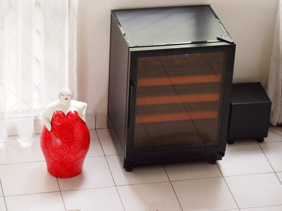 A beer storage cabinet (105 liters, height 84cm) next to a decorative red vase in a bright room with tiled floor and net curtains.