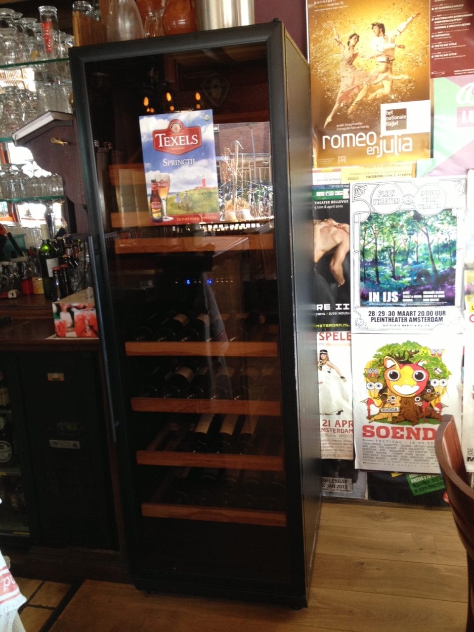 A high beer storage cabinet with glass front (105 liters, height 84 cm) filled with bottles, standing next to posters and advertisements in a bar setting.