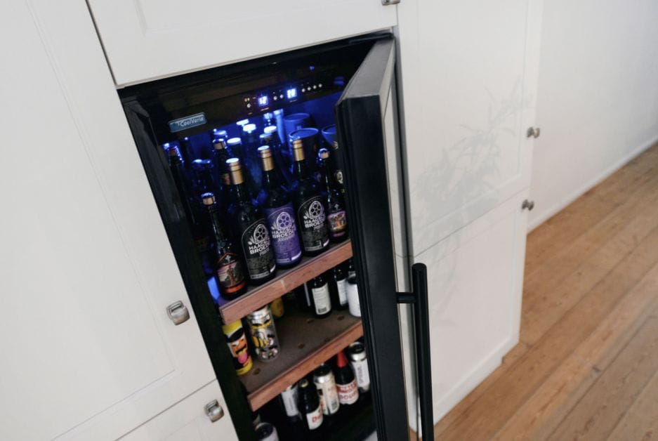 An open built-in beer climate cabinet (450 liters, height 180cm, multiple zones) full of various beer bottles, illuminated by blue interior lighting, in a modern kitchen.