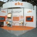 Messestand mit „abc“-Logo mit Displays, Informationstafeln und Sitzecke, in einer großzügigen Halle mit sichtbarer Deckenkonstruktion.