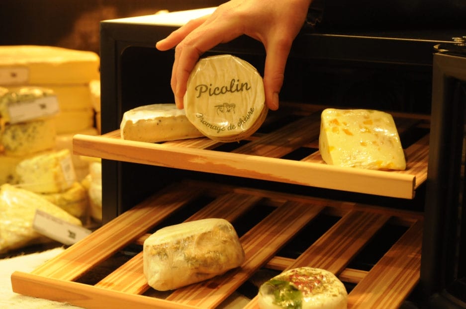 Une main plaçant une meule de fromage picolin dans l'enceinte climatique Fromage (25 litres + deux planches à fromage) sur une planche en bois remplie de différents types de fromages.