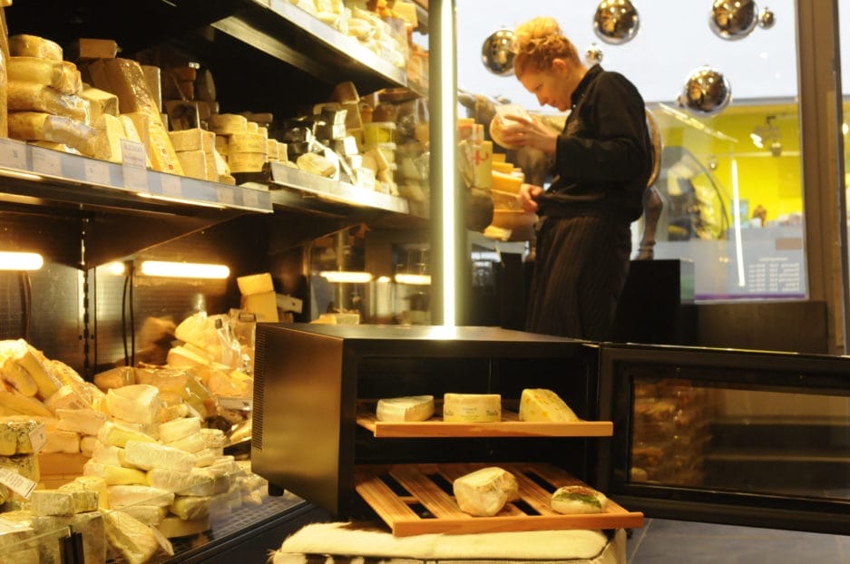 Eine Frau in schwarzer Schürze begutachtet Käse in einem gut sortierten Käseladen, im Vordergrund stehen verschiedene Käsesorten neben dem Käse-Klimaschrank (25 Liter + zwei Käsebretter).