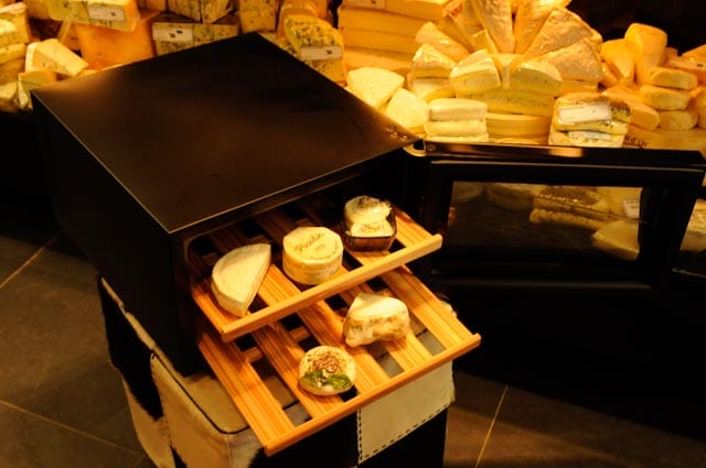 Une armoire climatique à fromages avec différents types de fromages sur des étagères en bois, entourée d'encore plus de types de fromages dans un magasin.