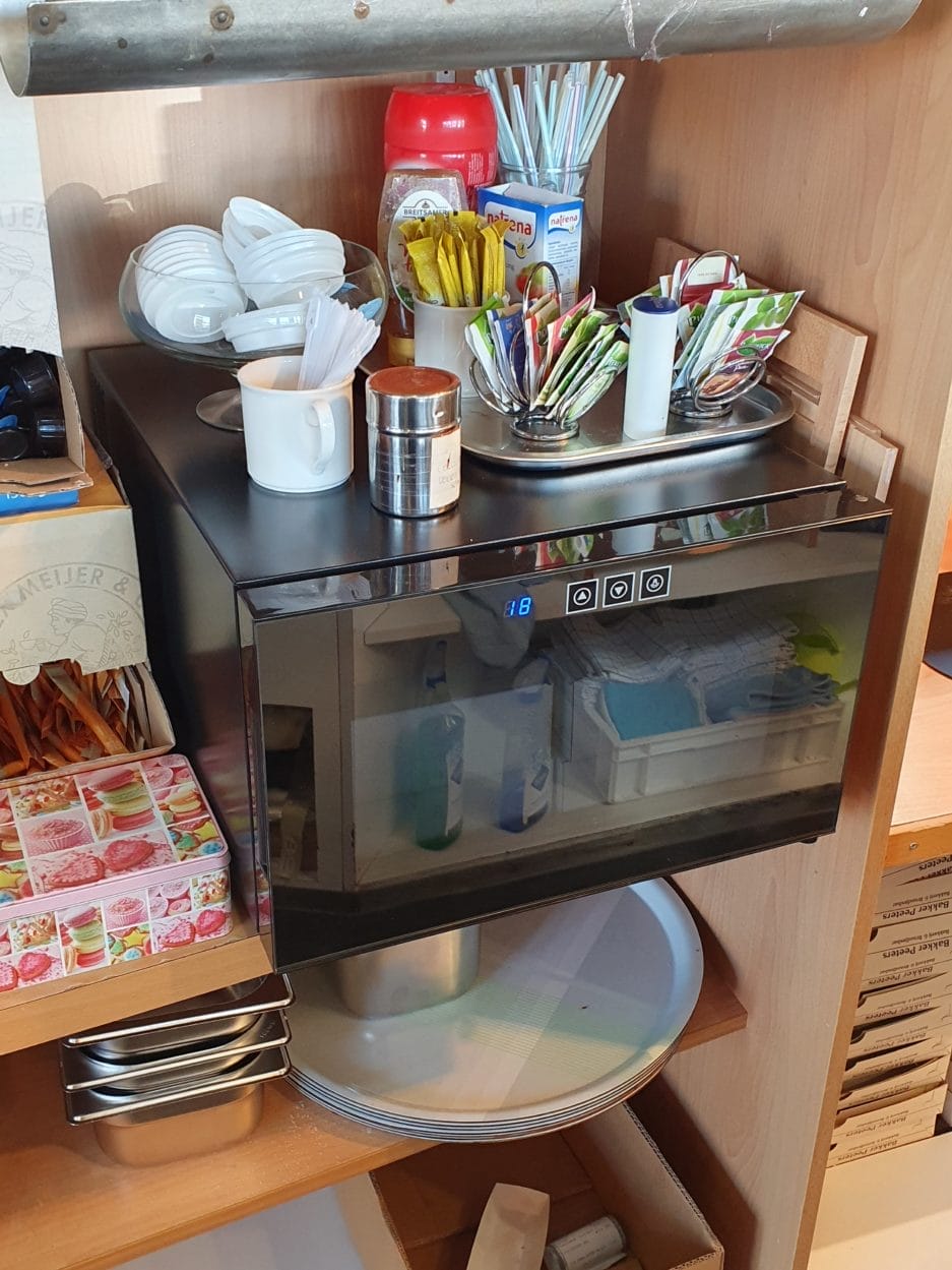 A messy countertop with a chocolate climate cabinet (25 liters), various kitchen utensils, plates and packets of spices.