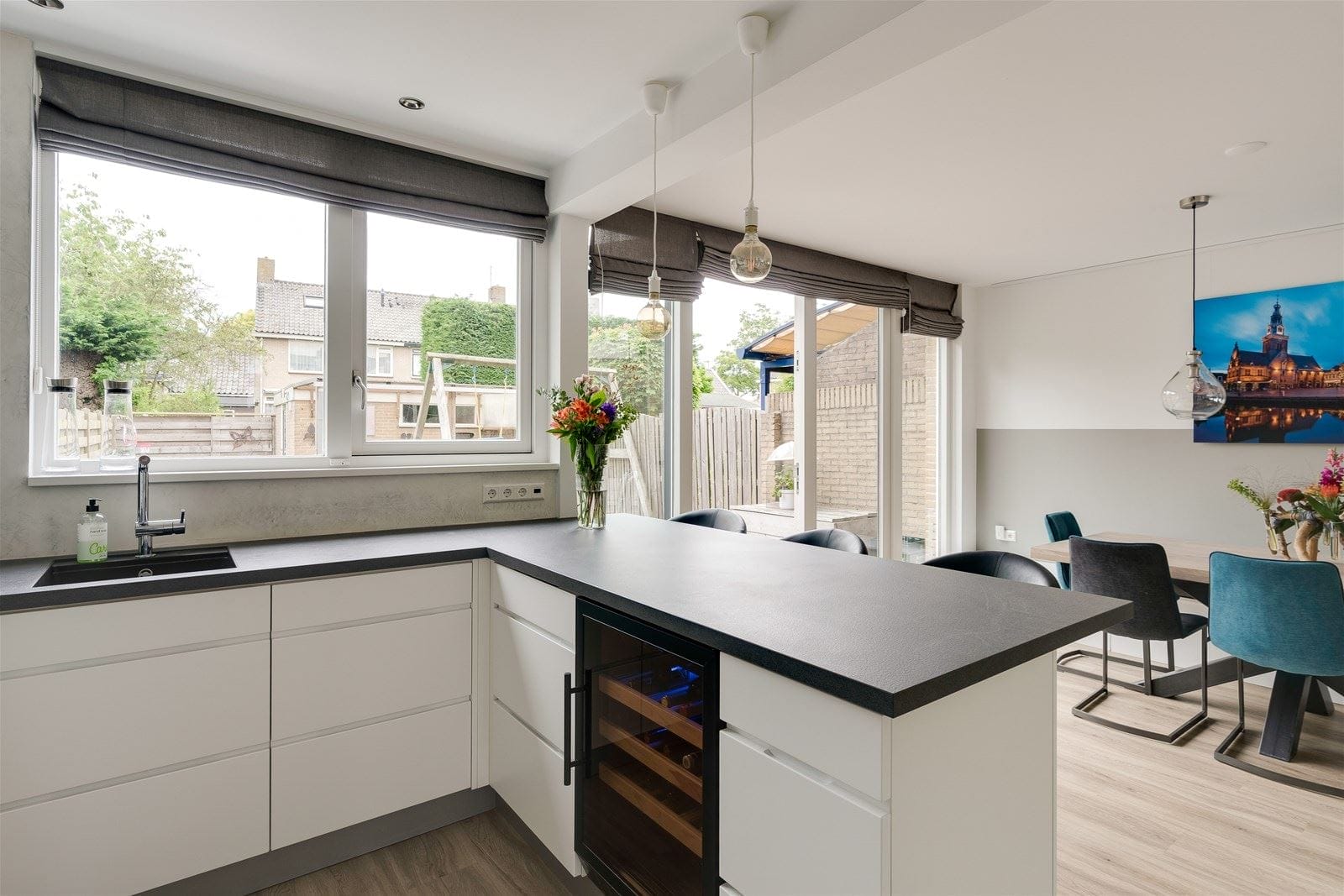 Modern kitchen with white cabinets and black worktop, equipped with a built-in wine cooler (40 bottles, multiple zones, 84cm height) and a dining area with blue chairs and colorful art on the wall.