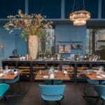A stylish dining area with teal chairs, a long wooden table set for a meal, an elegant lighting fixture above and a Wine cooler (40 bottles, multiple zones, 84 cm height) with glassware in the background.