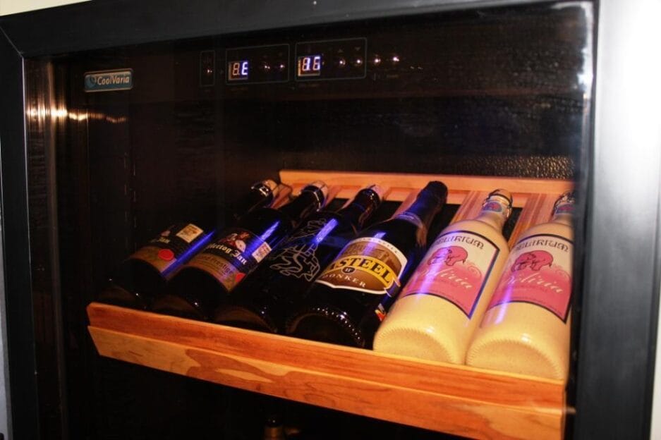Een bierkast met verschillende flessen bier uitgestald op een houten plateau, verlicht door een blauw licht aan de binnenkant dat de digitale temperatuur weergeeft.