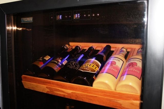 Ein Bierkühler mit mehreren Flaschen Bier auf einem Holzgestell, beleuchtet durch ein blaues Licht im Inneren, das digitale Temperaturwerte anzeigt.