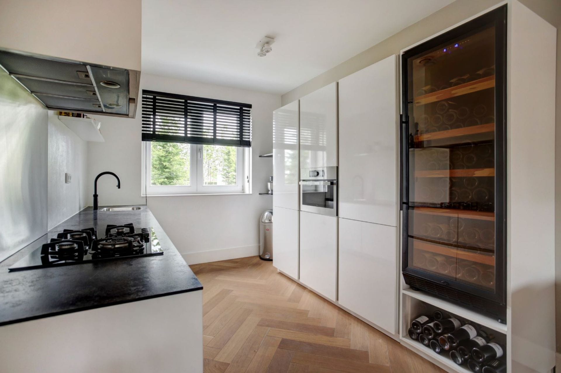 Modern keukeninterieur met zwart werkblad, witte kasten, een Wijnbewaarkast (200 flessen, één zone, 180cm hoogte, en een raam.