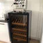 Une machine à expresso en acier inoxydable se dresse au sommet d'une armoire climatique à bière noire avec porte vitrée et étagères en bois dans une cuisine.