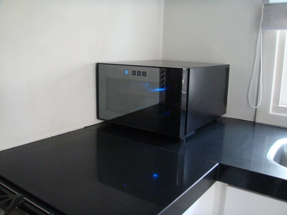A modern black Tea climate cabinet (25 liters) on a black granite countertop in a kitchen, with a blue clock showing 12:00.