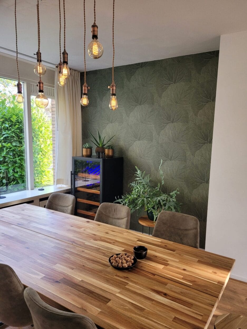 Moderne eetkamer met een grote houten tafel, hangende gloeilampen en behang met groenthema. op de achtergrond is een Bier klimaatkast (190 liter, hoogte 124cm, meerdere zones) zichtbaar.