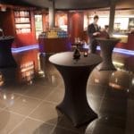 Espace bar moderne avec tables épurées et éclairage bleu, étagères remplies de boissons diverses et cave à vin (120 bouteilles, plusieurs zones, hauteur 164 cm) au travail.