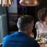 Zwei Menschen essen in einem Restaurant, im Mittelpunkt steht ein Mann von hinten, ihm gegenüber sitzt eine Frau, beide sitzen an einem Tisch mit Speisen und Getränken, während sie neben ihrem Weinklimaschrank (120 Flaschen, Mehrzonen, 164 cm Höhe) stehen.
