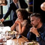 Een vrouw en een man dineren in een restaurant, glimlachend als ober die eten serveert terwijl hun Wijn klimaatkast (120 flessen, meerdere zones, 164 cm hoogte) vlakbij staat.