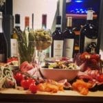 Eine Auswahl an Gourmetgerichten, darunter Aufschnitt, Käse und Oliven, umgeben von verschiedenen Weinkühlerflaschen (120 Flaschen, mehrere Zonen, 164 cm Höhe), auf einem Tisch.