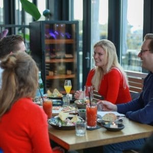 Vier mensen genieten samen van een maaltijd aan een restauranttafel gevuld met diverse gerechten en drankjes, in gesprek naast de Wijnklimaatkast (120 flessen, meerdere zones, 164cm hoogte).