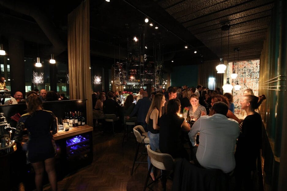A bustling bar scene at night with groups of people standing and sitting, chatting while enjoying a drink, illuminated by mood and hanging lighting.