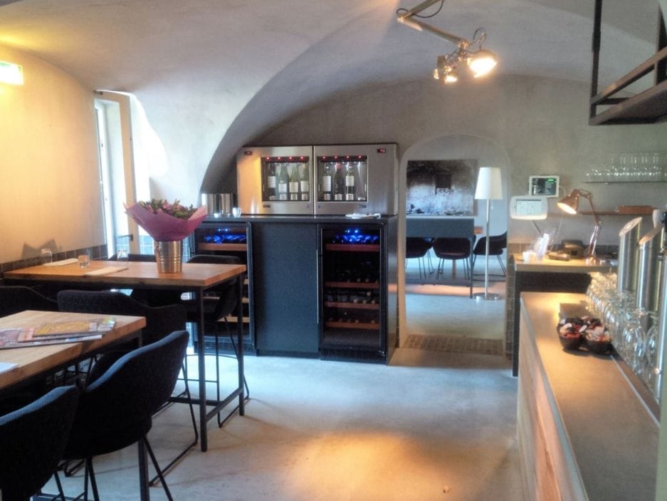 Interieur van een gezellig restaurant met houten tafels, barkrukken, wijnbewaarkasten ( max. 80 flessen, 124cm hoogte), en gewelfd plafond. Bovenop de bewaarkasten staat een "wijntap".