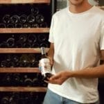 Homme souriant avec une bouteille Cave à vin (200 bouteilles, une zone, 180 cm de hauteur), debout devant un casier à vin rempli de diverses bouteilles.