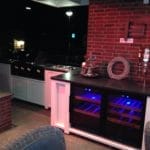 A modern home bar setting with an exposed brick wall, wine coolers, a storage shelf countertop stove and stylish bar stools in a warmly lit environment.