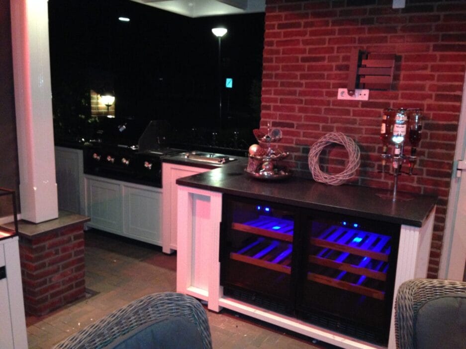A modern home bar setting with an exposed brick wall, wine coolers, a storage shelf countertop stove and stylish bar stools in a warmly lit environment.