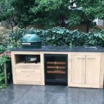 Meuble de cuisine extérieur avec étagère de rangement intégrée, armoires en bois et plan de travail en pierre, sur fond de verdure luxuriante.