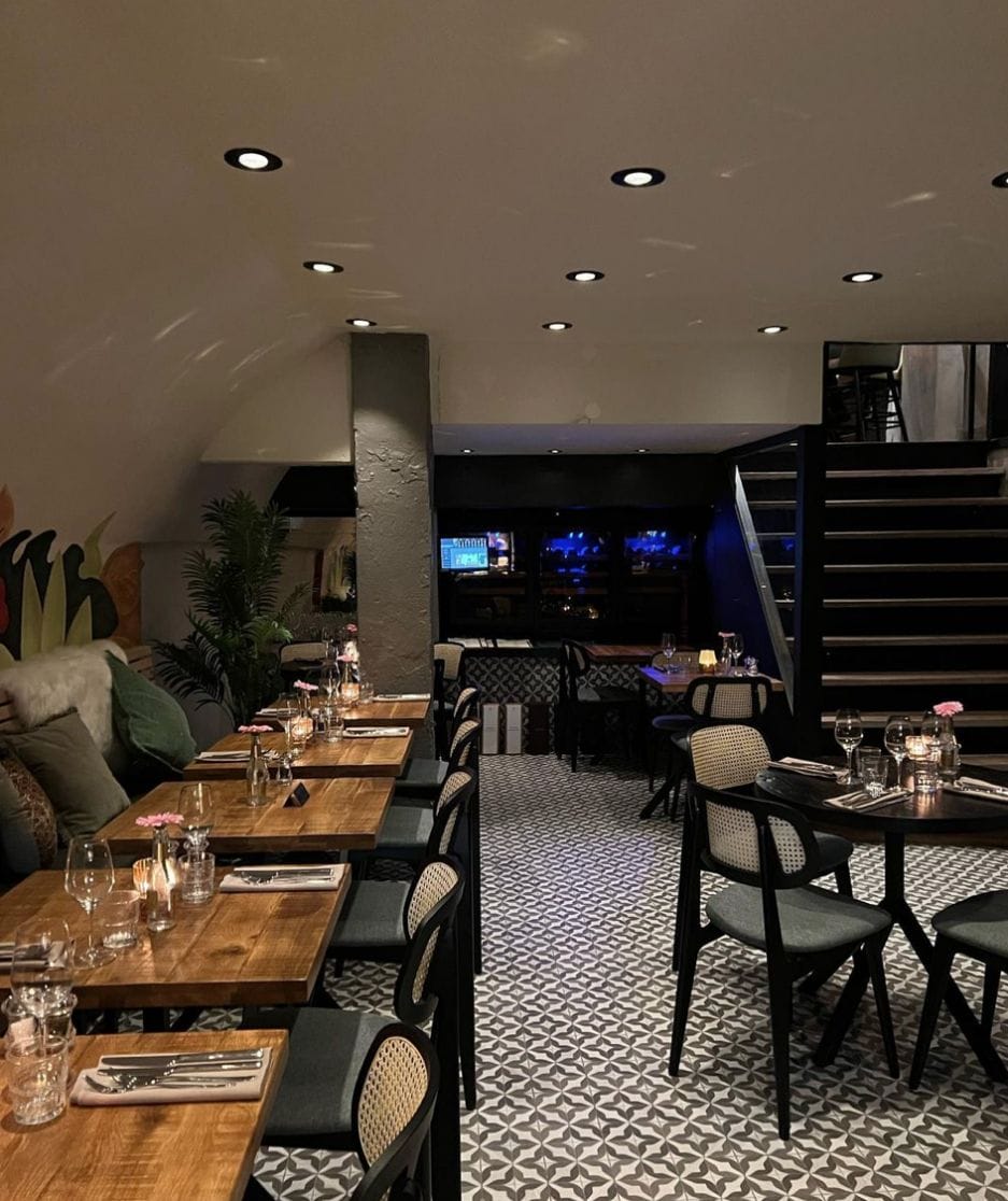 Luxe restaurantinterieur met Opslagplateau, netjes opgemaakte tafels met aankleding, groene planten en sfeerverlichting met een bar op de achtergrond.