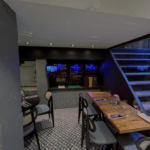 Interior of a modern restaurant with patterned floors, laid tables for dining, bar area, storage shelf in the background and a staircase on the right.