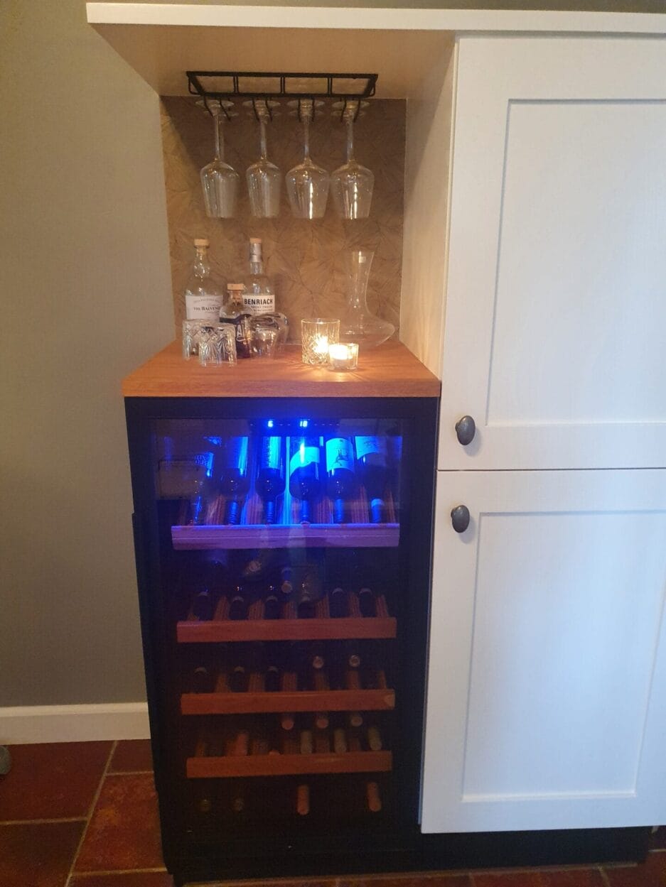 Een wijnbewaarkast gevuld met flessen, geplaatst onder een metalen rek met glazen en sterke drank, met sfeerverlichting.