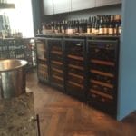 Intérieur de bar à vin moderne avec plusieurs réfrigérateurs à vin remplis de bouteilles, contre un mur bleu et un parquet.