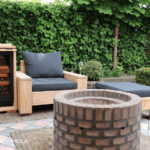 A cozy outdoor seating area with wooden benches, black cushions and a wine cooler, centered around a stone fire pit, surrounded by lush green hedges.