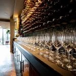 Des rangées de verres à vin vides bordent un bar, avec un présentoir mural de bouteilles de vin en arrière-plan d'un restaurant élégant.