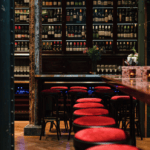 A cozy bar interior with a row of stools with a red top along a wooden counter, with a well-stocked shelf of bottles in the background.