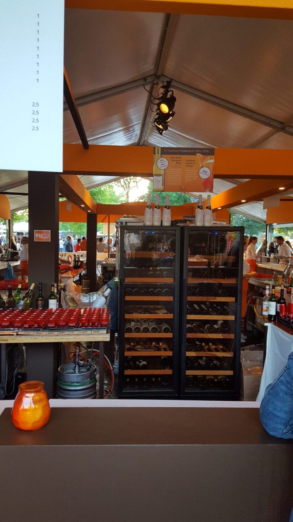 Outdoor wine bar with a glass refrigerator filled with wine bottles, shelves with drinks and customers in the background.