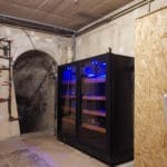 A modern wine cellar with a large black wine refrigerator with blue lighting, located in an underground stone room with vaulted ceilings and concrete floors.