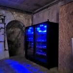 Des racks de serveurs éclairés en bleu dans une pièce souterraine sombre avec des murs en béton et une porte cintrée.
