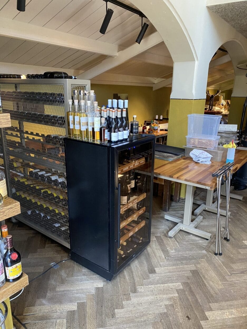 Een wijnbewaarkast gevuld met flessen, in een restaurant met houten meubilair en gebogen witte kolommen.