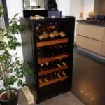 A wine refrigerator filled with bottles, placed in a modern kitchen next to a potted plant.