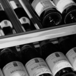 Rijen wijnflessen op houten planken, vanaf de zijkant gezien, met zichtbare etiketten in zwart-wit.