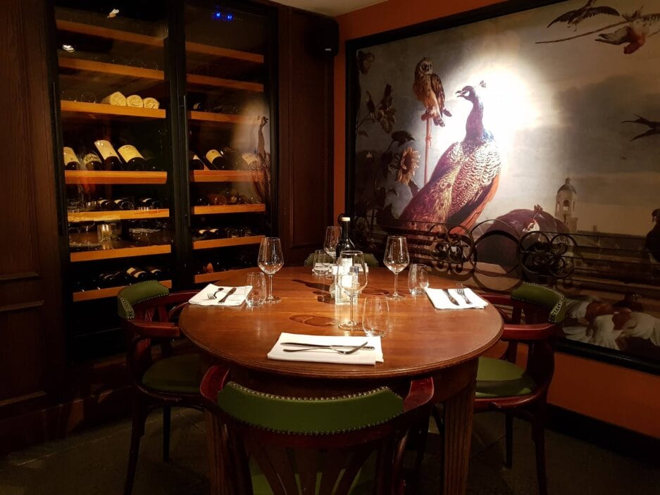 A round dining table for four in a cozy restaurant with a mural of a peacock on the wall and wine bottles on shelves.