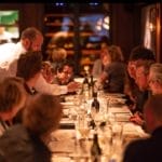 Un serveur sert du vin aux invités autour d’une longue table à manger dans un restaurant de luxe doté d’un éclairage d’ambiance.