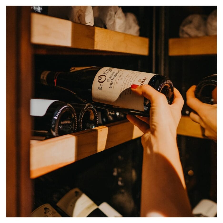 De hand van iemand die een fles wijn selecteert van een houten plank in een slecht verlichte kelder.