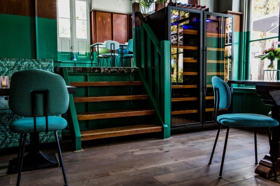 Interior of a stylish bar with teal chairs, green wooden steps and an illuminated wine fridge, combining a contemporary and cozy atmosphere.
