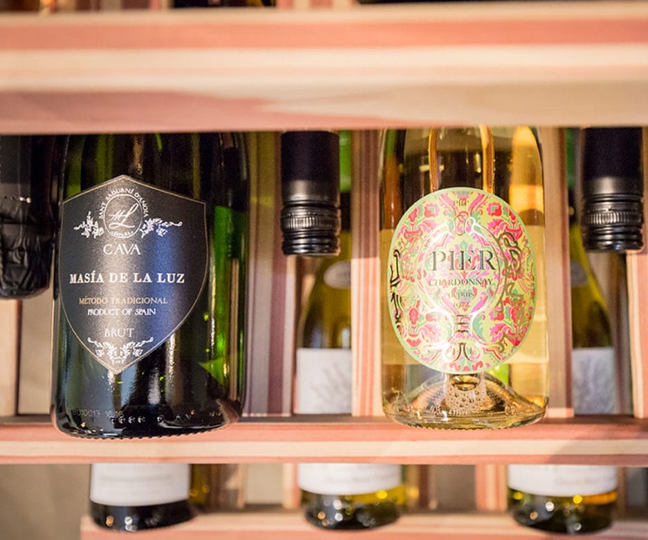 Flessen wijn en cava uitgestald op houten planken, met de nadruk op een kleurrijk etiket op een fles op de voorgrond.