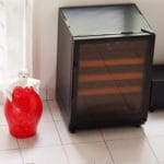 A modern wine cooler next to a red decorative vase in a room with white tile floors and net curtains.