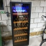 Ein Weinkühler mit Glastür, gefüllt mit Flaschen, beleuchtet von blauem Licht, steht neben einem Fahrrad in einer Garage mit einer Betonblockwand.